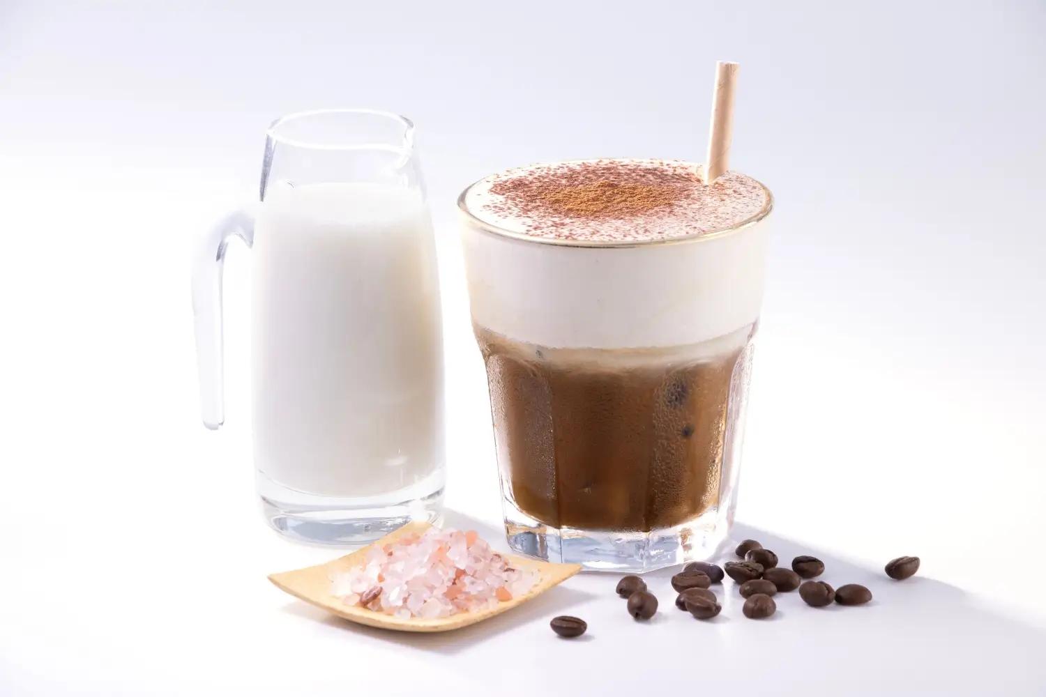 Sklenice mléka, ledová káva s pěnou a miska s růžovou solí vedle kávových zrn